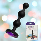 Image of the Doc Johnson Booty Bling Purple Silicone Anal Beads, showcasing three bulbous sections and a jeweled base, floating above a blurred, colorful background. To the right, there's Booty Bling packaging featuring the toy with its stunning purple-jeweled base.